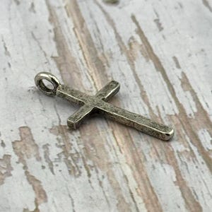 May include: A silver-colored cross pendant with a textured surface. The cross has a loop at the top for a chain. The background is a light-colored, weathered wood surface.