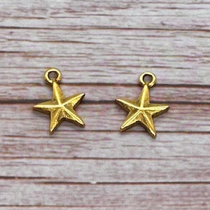 May include: Two gold-colored star charms with a loop at the top for hanging. The charms are five-pointed and have a textured surface. The background is a light brown wooden surface.