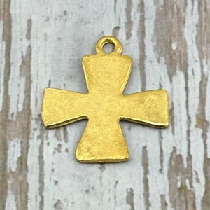 May include: A gold-colored metal cross pendant with a loop at the top for hanging. The cross has a simple, classic design with rounded edges and a slightly textured surface. The pendant is displayed against a light gray and white wooden background.