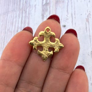 May include: A gold-coloured, textured metal charm in the shape of a cross. The charm has a small loop at the top for hanging and a detailed, ornate design. The charm is held in a hand with red nail varnish.