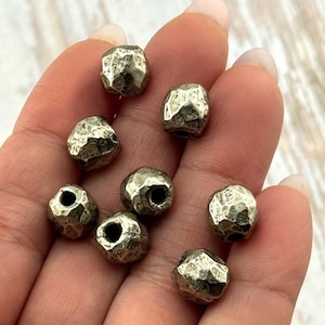 May include: A collection of silver-colored, faceted beads. Each bead has an irregular, textured surface and a hole running through the center. The beads are held in a hand, showcasing their metallic sheen and unique shape. Ideal for jewelry making.
