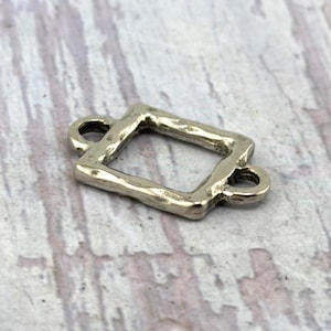 May include: A silver-toned metal connector charm featuring a rectangular open frame design with a textured, hammered surface. The piece includes two small circular loops, one on each of the shorter sides, intended for attaching to jewelry or craft projects.