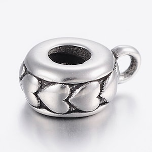May include: A silver-colored charm in the shape of a small teacup. The charm has a heart-shaped design around the base and a small handle. The top of the teacup has a large hole. The charm is made of metal.