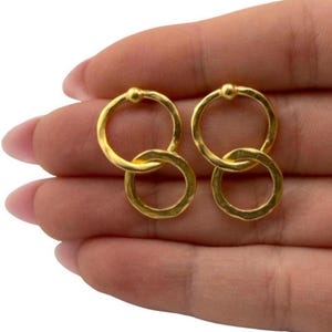 May include: A pair of gold-colored earrings. Each earring features two interlocking hammered rings. The top ring has a small ball at the top. The earrings are displayed on a light brown surface.