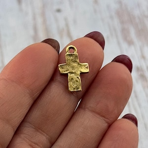 May include: A small, gold-colored cross pendant with a textured surface. The cross has a loop at the top for attaching to a chain or cord. The cross is a religious symbol.