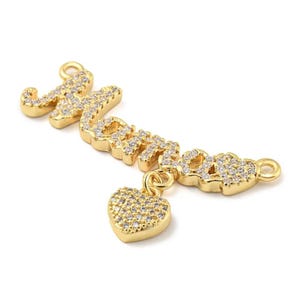 May include: Gold-toned pendant spelling "Mom" in a cursive font, encrusted with clear crystals. A small, crystal-covered heart charm dangles below the "Mom" pendant. The pendant has a loop at each end for attachment.