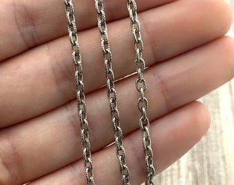 May include: Close-up of three silver-colored chain necklaces. The chains have an oval link design with a textured, rope-like pattern. The chains are held in a person's hand, with a light-colored background.