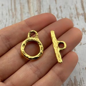 May include: Two gold-colored jewelry clasps. One is a textured ring with a loop at the top. The other is a textured bar with a loop at one end. The clasps are likely for necklaces or bracelets.