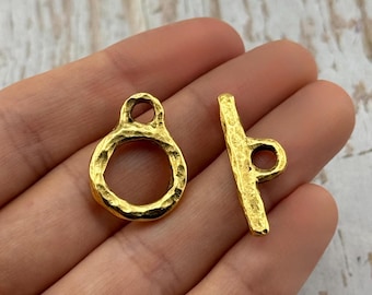 May include: Two gold-colored jewelry clasps. One is a textured ring with a loop at the top. The other is a textured bar with a loop at one end. The clasps are likely for necklaces or bracelets.