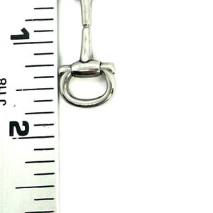 May include: A silver-colored horse bit, approximately 3 inches in length, with a central bar and rounded rings on each end. The bit is positioned vertically against a ruler.