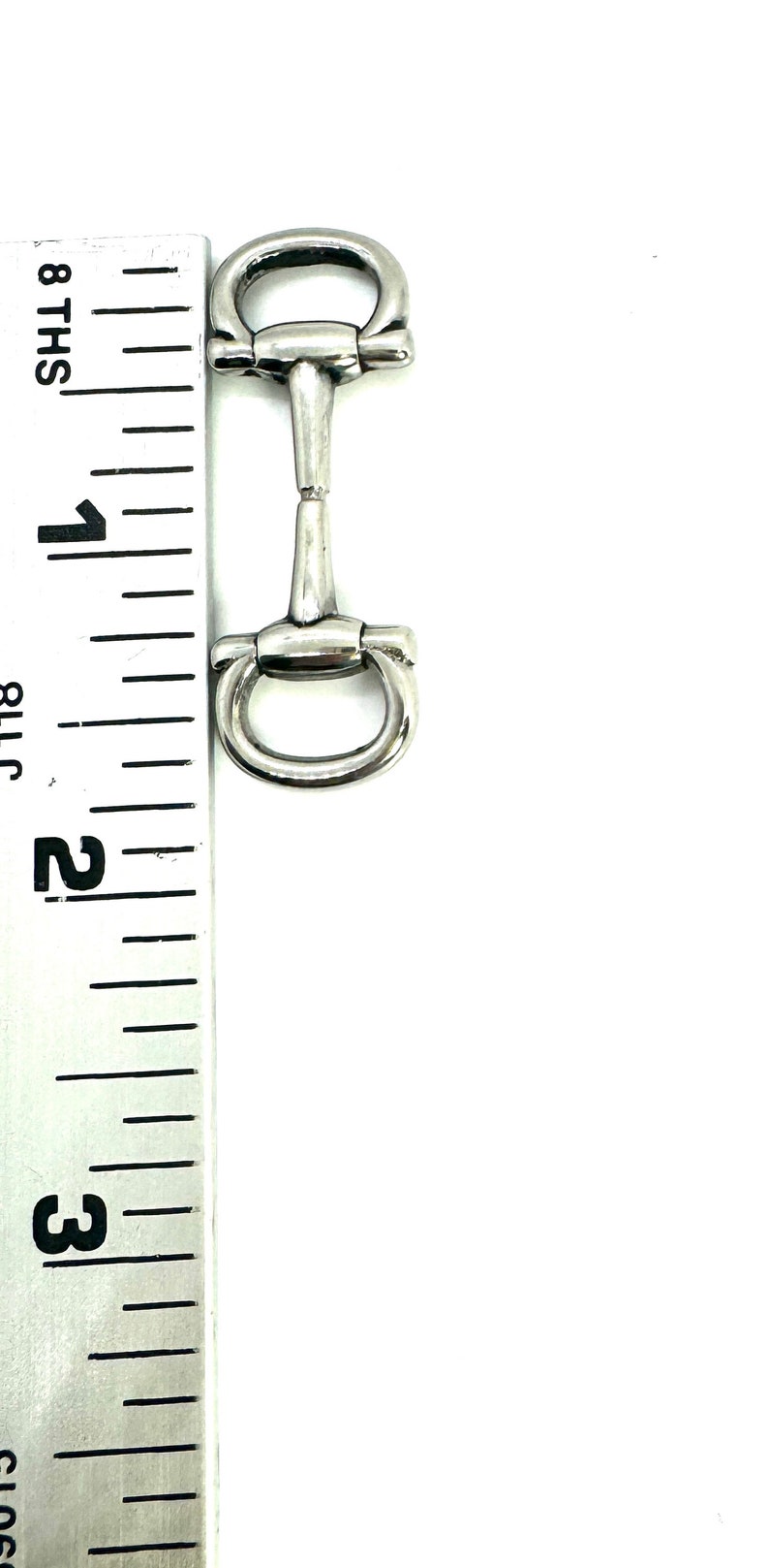 May include: A silver-colored horse bit, approximately 3 inches in length, with a central bar and rounded rings on each end. The bit is positioned vertically against a ruler.