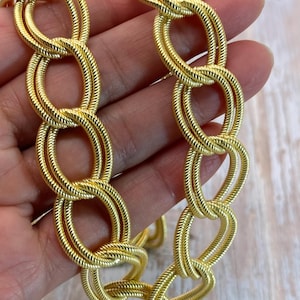 May include: A gold-colored chain necklace with a textured, rope-like design. The chain is composed of interconnected oval links, creating a bold and eye-catching accessory. The necklace is held against a light wood-grain surface.