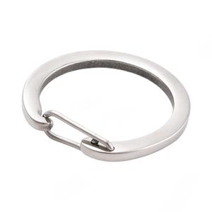 May include: A silver-colored, circular keychain with a hinged clasp. The keychain is made of a smooth, metallic material and has a simple, minimalist design. The clasp is a small, straight bar that connects to the ring.