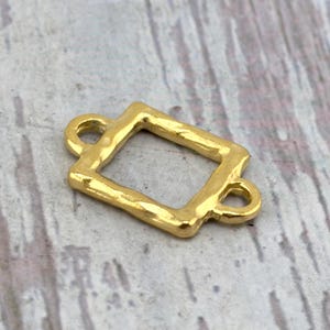 May include: A gold-colored metal connector charm featuring a rectangular frame with a slightly irregular, hammered texture. The piece includes two small circular loops, one on each of the shorter sides, designed for attaching to jewelry or craft projects.