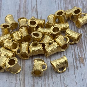 Puede incluir: Una colecci&oacute;n de peque&ntilde;os dijes de metal dorado con forma de jarra de cerveza. Cada dije tiene una superficie texturizada y un asa peque&ntilde;a. Los dijes est&aacute;n esparcidos sobre una superficie de madera clara.