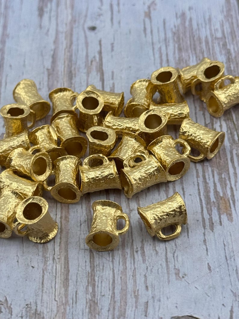 Puede incluir: Una colecci&oacute;n de peque&ntilde;os dijes de metal dorado con forma de jarra de cerveza. Cada dije tiene una superficie texturizada y un asa peque&ntilde;a. Los dijes est&aacute;n esparcidos sobre una superficie de madera clara.