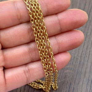 May include: Close-up of several strands of a gold-colored chain. The chains are made of small, oval-shaped links, creating a delicate and intricate design. The chains are held in a hand, with a wooden surface in the background.
