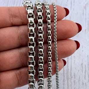 May include: Close-up of four silver-colored box chain necklaces of varying widths. The chains are held against a light-colored wooden surface. The chains are made of interlocking rectangular links, creating a sleek, modern look. The chains are displayed in different sizes.