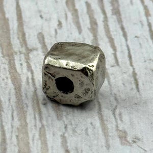 May include: A silver-colored, cube-shaped bead with a slightly irregular surface. The bead has a hole drilled through the center, suitable for stringing. The bead's design suggests it could be used in jewelry making or other craft projects.