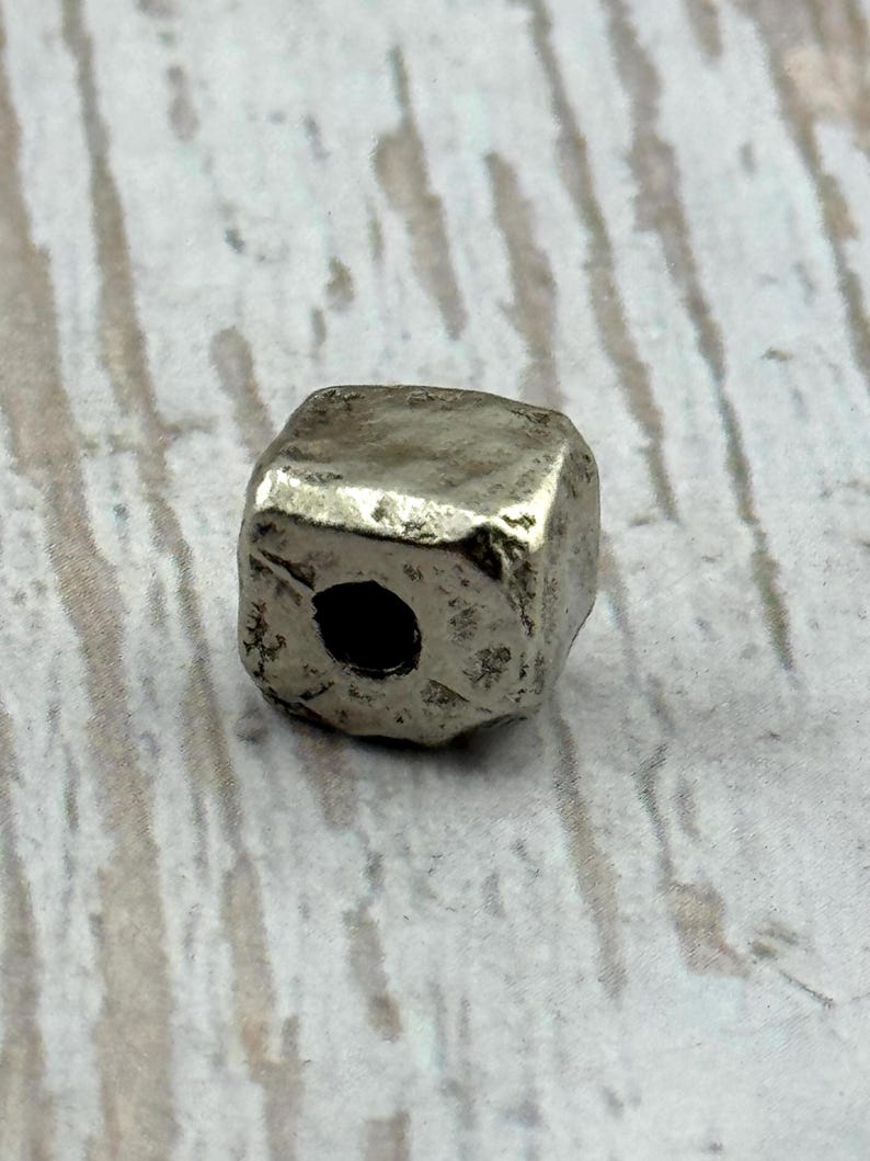 May include: A silver-colored, cube-shaped bead with a slightly irregular surface. The bead has a hole drilled through the center, suitable for stringing. The bead's design suggests it could be used in jewelry making or other craft projects.