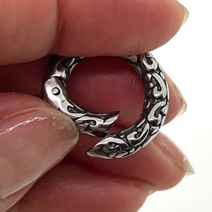 May include: A silver-toned metal clasp with an intricate, ornate design. The clasp is open and ready to be attached to a chain or other jewelry.