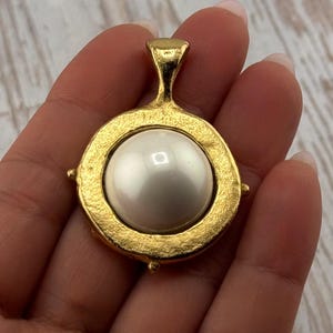 May include: A gold-toned pendant featuring a large, lustrous, white pearl. The pearl is set within a textured, circular frame with small decorative accents. The pendant has a loop for a chain. Jewelry.