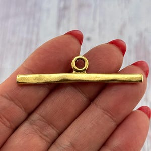 May include: A gold-colored bar with a loop at the top, held in a hand with red nail polish. The bar is rectangular with rounded edges and a slightly textured surface. The loop is circular and attached to the center of the bar.