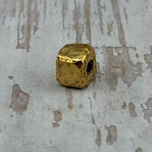May include: A small, gold-colored bead with a cube-like shape. The bead has a rough, textured surface and a hole drilled through one side. The bead is likely used for jewelry making or other crafts.