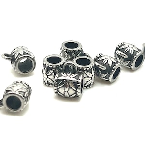 May include: A collection of silver-colored metal beads. Each bead is cylindrical with a floral design and a small loop. The beads are scattered on a white surface, showcasing their intricate details and reflective surfaces.