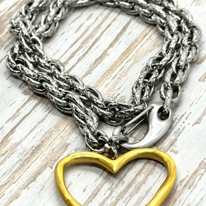 May include: Silver-toned chain necklace with a gold-colored heart pendant. The necklace features a thick, textured chain and a lobster clasp. The heart pendant is a simple, open design.