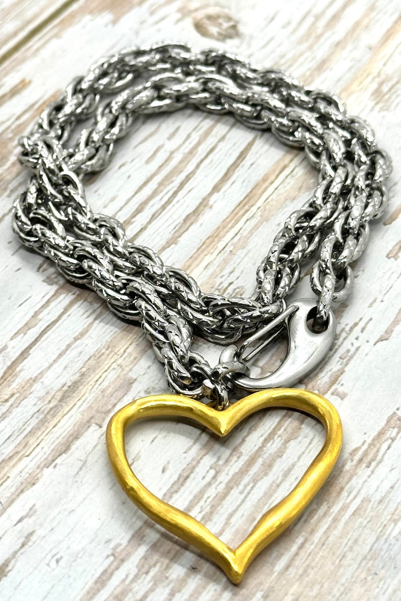 May include: Silver-toned chain necklace with a gold-colored heart pendant. The necklace features a thick, textured chain and a lobster clasp. The heart pendant is a simple, open design.