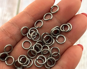 May include: A collection of small, silver-colored jump rings with a textured, twisted rope design. The rings are open and ready for use in jewelry making or other crafts. The rings are held in a hand with red nail polish.