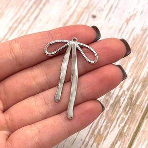 Puede incluir: Un dije de lazo plateado con un lazo en la parte superior y dos colgantes alargados y curvos. El dije se sostiene en una mano sobre un fondo claro de vetas de madera. Adecuado para la fabricaci&oacute;n de joyas.