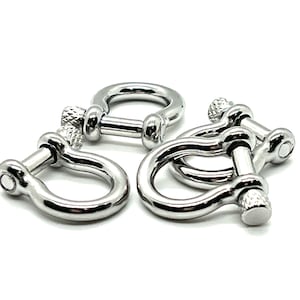 May include: Close-up of four shiny, silver-colored D-shackle fasteners. Each shackle has a U-shaped body with a threaded pin closure. The fasteners are arranged on a white background, showcasing their polished metal finish.