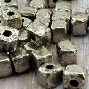May include: A collection of silver-colored, cube-shaped beads with a textured, hammered appearance. Each bead has a hole running through the center, suitable for stringing. The beads are scattered, showcasing their irregular surfaces.