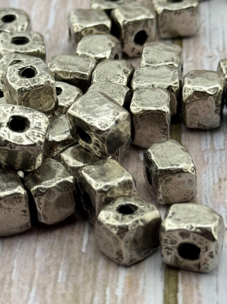 May include: A collection of silver-colored, cube-shaped beads with a textured, hammered appearance. Each bead has a hole running through the center, suitable for stringing. The beads are scattered, showcasing their irregular surfaces.