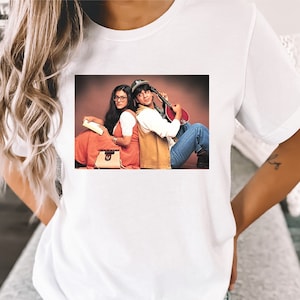 May include: White t-shirt with a printed image of a man and woman sitting back-to-back. The man is wearing a brown jacket and blue jeans. The woman is wearing a red dress and holding a book.
