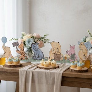 May include: A wooden table is decorated with Winnie the Pooh character cutouts, cupcakes, honey jars, and floral arrangements. The scene includes Pooh, Tigger, Eeyore, Piglet, and other characters. The cupcakes are on wooden slices, and the flowers are in pastel colors.