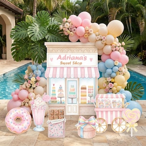May include: A party backdrop featuring a storefront sign reading Adriana's Sweet Shop, surrounded by a balloon arch in pink, blue, and cream. Cutouts include a frosted donut, a milkshake, a chocolate bar, a gift box, and a candy cart.