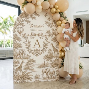 May include: An arched backdrop for a christening featuring a beige and brown toile pattern with illustrations of elephants, giraffes, and a hot air balloon, centered with the text Alexander's Christening and the letter A, decorated with beige and gold balloons.