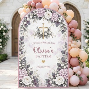 May include: An arched baptism sign featuring a gold cross, floral illustrations in white and purple, and the text "ON THIS SPECIAL DAY", "Olivia's", "BAPTISM", and "05.06.2026". The sign is framed by a large balloon arch in shades of pink and peach.