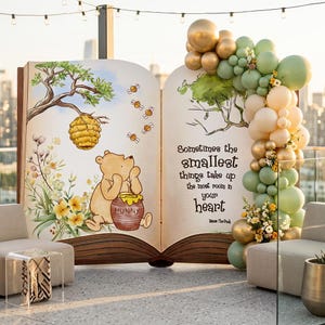 May include: A large, open book-shaped display featuring a Winnie the Pooh illustration with a quote. The display is decorated with a balloon arch in gold, green, and cream colors, and is set up outdoors.