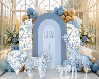 May include: A blue arched backdrop featuring the name LIAM and the number 1, surrounded by blue, white, and gold balloons. Decorative cutouts include a tiger, a giraffe, a hot air balloon, and a bow, all with a blue and white toile pattern.