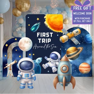 Könnte beinhalten: Ein Party-Hintergrundset zum Thema Weltraum mit einem dunkelblauen rechteckigen Schild mit dem Text FIRST TRIP Around the Sun, einer gelben Sonne, einem Kometen, einem Astronauten, einer Rakete, einem Teleskop, einem Mond und einem Planeten.
