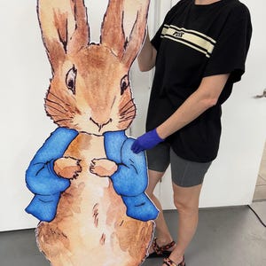 May include: A large cardboard cutout of Peter Rabbit from the children's book by Beatrix Potter. The cutout is brown with a blue jacket and is standing next to a person wearing a black shirt and shorts.