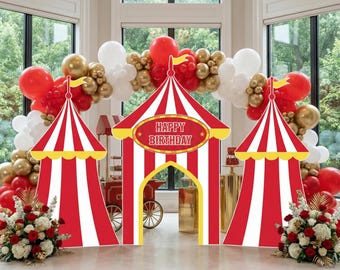 May include: A circus-themed party decoration set featuring three red and white striped tent cutouts, with the center tent displaying the text "HAPPY BIRTHDAY" in a yellow oval. The display is framed by an arch of red, white, and gold balloons.