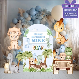 Op de afbeelding: Een dinosaurus thema verjaardagsfeest decoratie set. Inclusief een bord met "Happy Birthday! MIKE ROAR", dinosaurus uitsparingen en een jeep vol dinosaurussen. De achtergrond heeft ballonnen en bladeren.