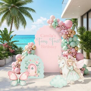 May include: A large pink arched backdrop with the text Camila's Fairy First Birthday, decorated with a balloon garland in shades of pink, mint green, and gold. Includes a mint green fairy door cutout, a pink butterfly, and a fairy with a unicorn.