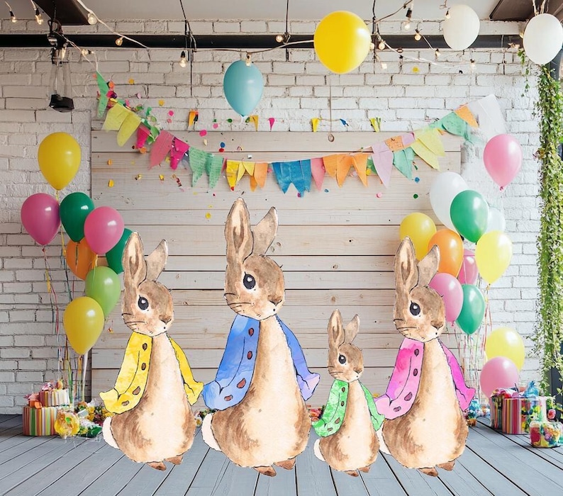 May include: Four Peter Rabbit characters in different colored sweaters stand in front of a brick wall decorated with colorful flags and balloons. The characters are wearing blue, yellow, pink, and green sweaters.