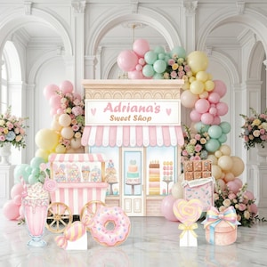 May include: A backdrop featuring a candy shop facade labeled Adriana's Sweet Shop, decorated with pastel balloon arches, a milkshake, a donut, a chocolate bar, and a gift box, all set against a white architectural interior with floral arrangements.